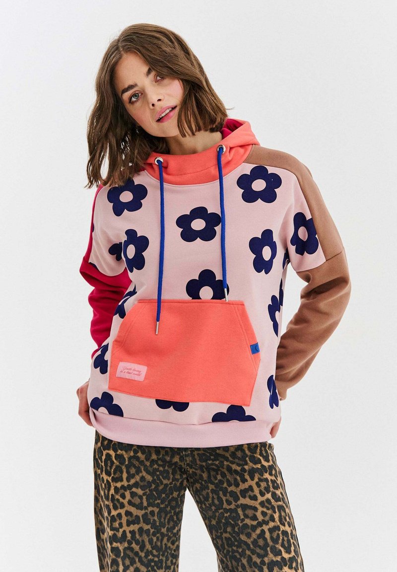 Ružová mikina s kapucňou a námorníckym kvetinovým vzorom, s korálkovou vreckom, modrými šnúrkami a kontrastnými hnedými rukávmi. Nosená s nohavicami s leopardím vzorom.