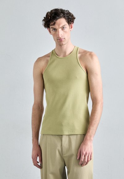 Grünes geripptes Tanktop mit hohem Kragen und breiteren Trägern, kombiniert mit beigen Hosen, vor einem grauen Hintergrund präsentiert.