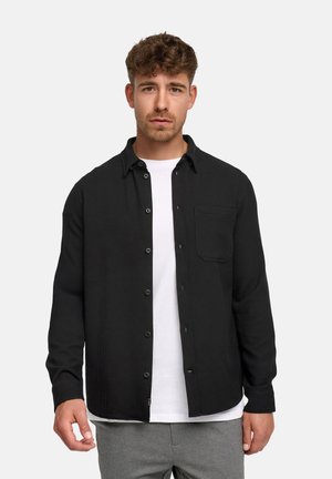 Mand med kort krøllet hår iført en sort knappet skjorte over en hvid t-shirt og grå bukser mod en ensfarvet lys baggrund.