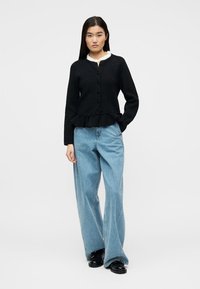 Γυναίκα που φοράει μαύρο καρό ζακέτα με κουμπιά και βολάν, λευκό πουκάμισο με βολάν, φαρδιά ανοιχτό μπλε τζιν παντελόνια και μαύρα παπούτσια, στέκεται με το χέρι στην τσέπη.