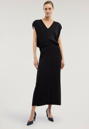 Frau trägt ein ärmelloses schwarzes Langkleid mit V-Ausschnitt und Schulterpolstern, kombiniert mit schwarzen High Heels, stehend vor einem neutralen Hintergrund.