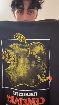 Melns T-krekls ar lielu dzeltena ābola grafiku, kurā redzami divi sarkani un balti svītraini tārpi. Teksts lasāms "Skolotāja mājdzīvnieku kapsēta" sarkanā krāsā.