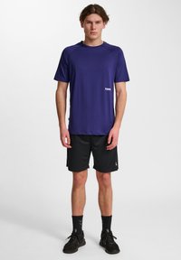 Maglietta atletica corta viola con colletto tondo, abbinata a pantaloni neri. Presenta un logo sul lato sinistro della maglietta.