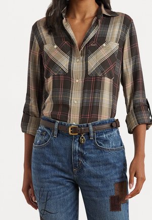 Woman wearing a brown plaid button-up shirt with rolled sleeves, tucked into blue jeans with patch details and a brown belt with a small charm.