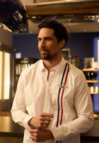 Homme aux cheveux bouclés sombres et à la barbe, portant une chemise blanche à boutons avec des rayures verticales rouges et bleues et un emblème ailé, se tenant à l'intérieur.