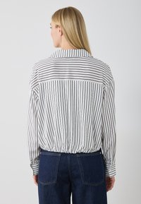 Hvit skjorte med svarte vertikale striper, med kragehals og elastisk rynket kant, sammen med mørkeblå denimbukser.