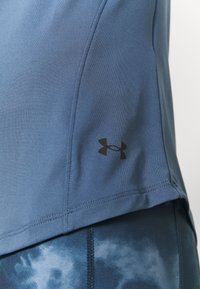 Blauses Sportshirt aus glattem Material mit einem Seitennähtedesign und einem schwarzen Under Armour-Logo in der Nähe des Saums, kombiniert mit gemusterten Leggings.
