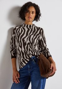 Pull en motif zèbre dans des tons de beige et marron. Doté d'une encolure haute et d'une coupe ample, associé à un jean bleu et un sac en suède marron.