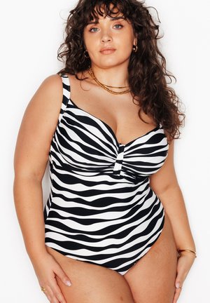 Donna con capelli ricci che indossa un costume intero a righe zebrate bianche e nere e gioielli d’oro, in piedi davanti a uno sfondo bianco.