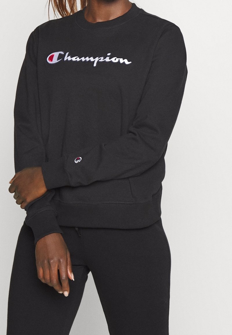 Sweat-shirt noir à col rond, logo Champion brodé en blanc et rouge sur la poitrine, poignets et ourlet côtelés.