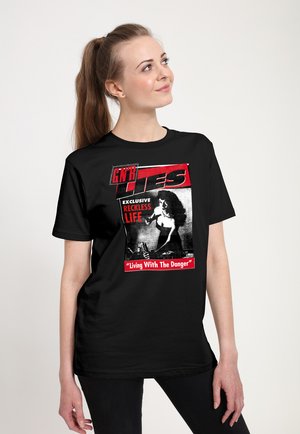 Giovane donna con capelli castani legati in una coda di cavallo indossa una maglietta nera con una grafica rossa e bianca con scritto "GNR Lies," "Exclusive Reckless Life," e "Living With The Danger."