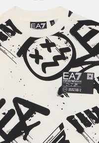 Krämfärgad t-shirt i bomull med abstrakta svarta grafiska mönster och en märkeslapp med QR-kod vid fållen.