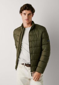 Doudoune vert olive avec des sections matelassées et un col haut, portée par-dessus un pull gris clair. Comprend une fermeture éclair et des poches latérales.