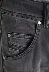 Svarta denimjeans med en närbild på fickdetaljerna, som har metallknappar och en tygetikett. Mjuk textur med en lätt blekning.