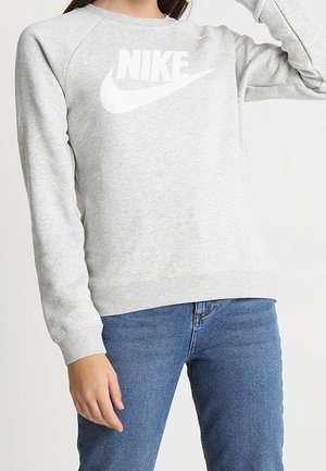 Kvinde iført en lysegrå Nike-sweatshirt med hvidt logo og blå jeans, stående med den ene arm hævet.