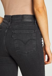 Jeans de mezclilla negra con una cintura alta, bolsillos traseros distintivos con un patrón de costura curvado y una etiqueta de marca de cuero negra en la parte trasera.