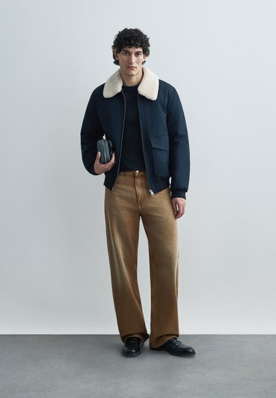 Mornarsko modra bomber jakna z ovratnikom iz kremastega krzna, črno srajco, širokimi rjavimi kavbojkami in črnimi čevlji. V roki drži majhno temno sivo torbico.