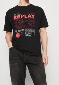 Černé tričko s červeným a bílým nápisem "REPLAY" opakovaným, grafika červené planety, souřadnice Země a Marsu, nošeno s tmavými vybledlými džínami.