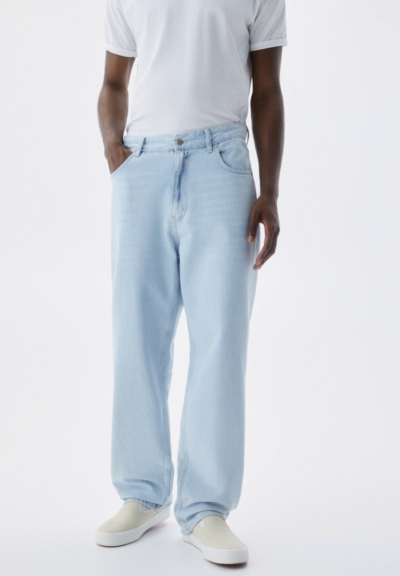 Jeans de mezclilla de un azul claro, de corte holgado, con cierre de botón y cremallera, bolsillos delanteros y una ligera textura desgastada, combinados con zapatos blancos.