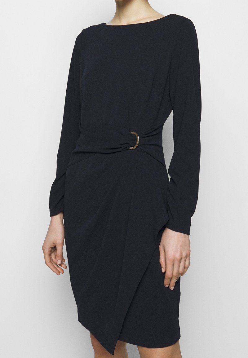 Femme portant une robe noire à manches longues avec un col rond et une taille froncée maintenue par un petit anneau métallique.