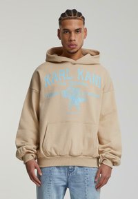 Felpa beige con cappuccio, testo blu e grafica di un'aquila, dotata di tasca frontale a kangaroo e polsini e orlo a coste.