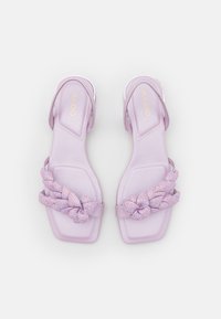 Sandales lavande avec une semelle plate, dotées d'une sangle tressée ornée d'accents scintillants et d'un design ouvert à l'arrière. Fabriquées en matériau synthétique.