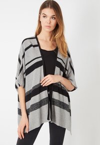 Una mujer con cabello largo lleva un cárdigan gris holgado con rayas negras sobre una camiseta negra y pantalones negros, de pie contra un fondo liso.