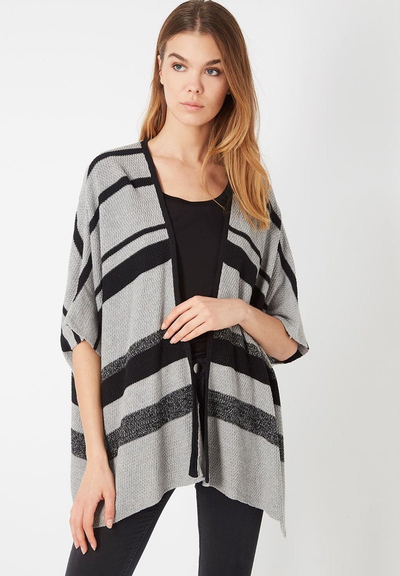 Una mujer con cabello largo lleva un cárdigan gris holgado con rayas negras sobre una camiseta negra y pantalones negros, de pie contra un fondo liso.