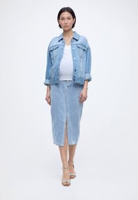 Femeie însărcinată purtând o jachetă din denim albastru deschis și o fustă midi asortată cu o despicătură frontală, top alb și tocuri argintii, stând în fața unui fundal alb.