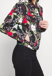 Giacca di jeans nera con motivo floreale, decorata con foglie verdi e fiori rossi, dotata di colletto, maniche lunghe e chiusura frontale con bottoni.
