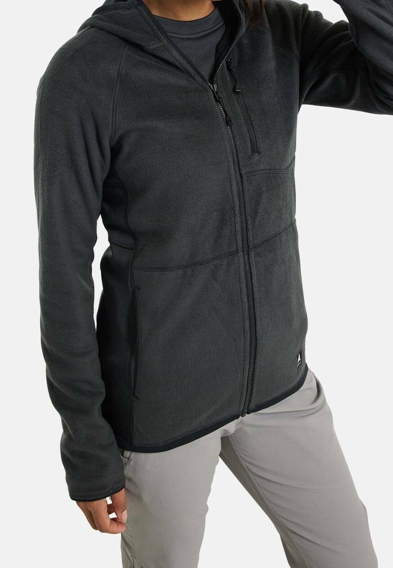 Personne portant une veste polaire zippée gris foncé avec capuche et un pantalon gris clair sur un fond uni.