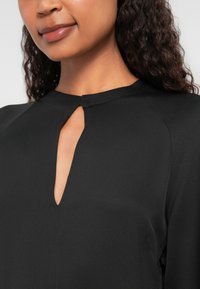 Blusa nera con scollatura a occhio di bottone, dalla textura morbida e un leggero lucido. Presenta dettagli di cucitura discreti sulle spalle.