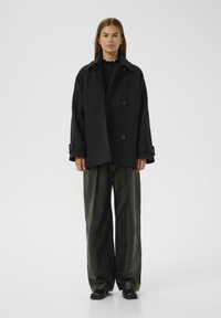Black wool blend double-breasted coat with large buttons, wide lapels, and rolled sleeves, paired with loose-fitting dark leather trousers.