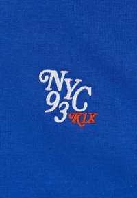 Blått stoff med brodert tekst: "NYC 93" i hvitt og "K1X" i oransje, med en glatt, teksturert overflate.