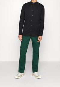 Camisa negra de manga larga con cuello alto y cierre de botones, combinada con pantalones verdes y zapatillas blancas, sobre un fondo liso.