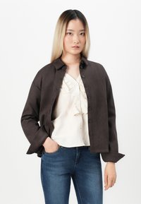 Brown button-up jacket with a relaxed fit, worn over a cream blouse featuring ruffles, paired with blue jeans. Soft texture and lightweight design.
