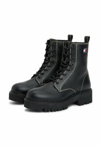 Botas de cuero negras con cordones, suelas gruesas de taco y costuras blancas, y un pequeño logo de Tommy Jeans en el lado cerca de la parte superior.