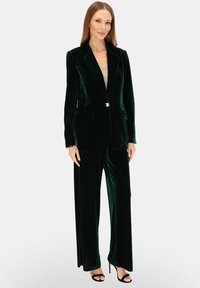 Zielony aksamitny garnitur z dopasowaną marynarką i spodniami z szerokimi nogawkami. Marynarka ma dwie przednie kieszenie oraz dekoracyjny guzik.