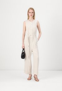 Combinaison en lin beige avec une fermeture boutonnée à l'avant, ceinture à la taille, silhouette à jambes larges, assortie à un petit sac à main noir et des sandales à lanières couleur tan.