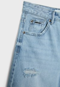 Jean en denim bleu clair avec une taille haute, des détails déchirés avec des bords effilochés et des accents en métal cuivré sur les poches.