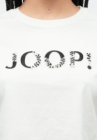 Άσπρο βαμβακερό μπλουζάκι με το κείμενο «JOOP!» σε μαύρη κεντητική, διακοσμημένο με μαύρες και άσπρες χάντρες για υφή και λεπτομέρεια.