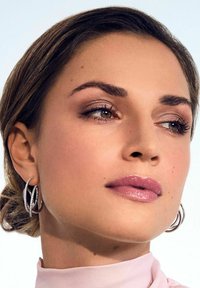 Orecchini a cerchio argentati, rossetto rosa morbido, trucco occhi discreto e blusa beige chiaro. Capelli lisci con morbide onde. Sfondo neutro.