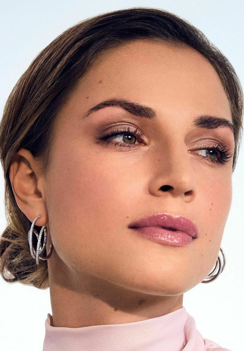 Orecchini a cerchio argentati, rossetto rosa morbido, trucco occhi discreto e blusa beige chiaro. Capelli lisci con morbide onde. Sfondo neutro.