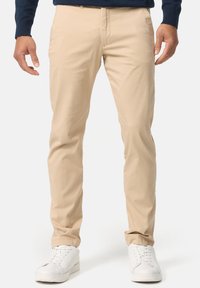 Khaki chinos vyrobené z hladké bavlněné látky, mají slim fit, standardní kapsy a jsou kombinovány s bílými teniskami pro volnočasový vzhled.