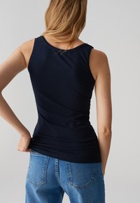 Femme aux cheveux blonds portant un top sans manches bleu marine et un jean bleu, de dos, tenant l'ourlet inférieur du top d'une main.