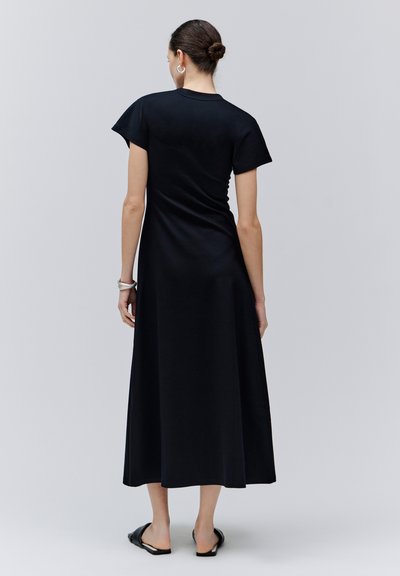 Femme aux cheveux attachés en chignon, portant une robe midi noire à manches courtes et des chaussures noires sans lacets, debout de dos face au spectateur.