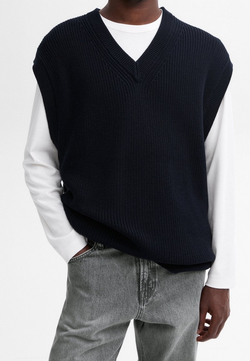Marineblauer Strickpullover mit V-Ausschnitt, getragen über einem weißen Langarmshirt. Verfügt über eine lockere Passform und gerippten Textur. Kombiniert mit grauen Jeans.