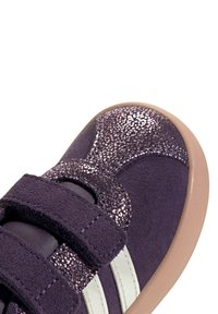 Zapatilla púrpura de material de ante, con acentos brillantes, correas de velcro y rayas blancas. Punta redondeada y suela de goma de color claro.