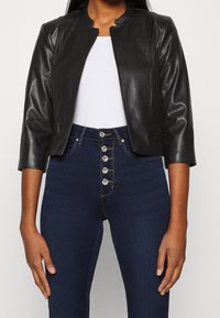 Veste en cuir noir raccourcie avec un col haut et des manches trois-quarts, associée à un jean taille haute en denim foncé avec des boutons décoratifs.