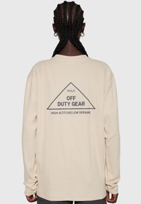 Beige långärmad topp i strukturerat tyg. Baksidan pryds av en triangelgrafik med texten: "HALO OFF DUTY GEAR HIGH ALTITUDE LOW OPENING."